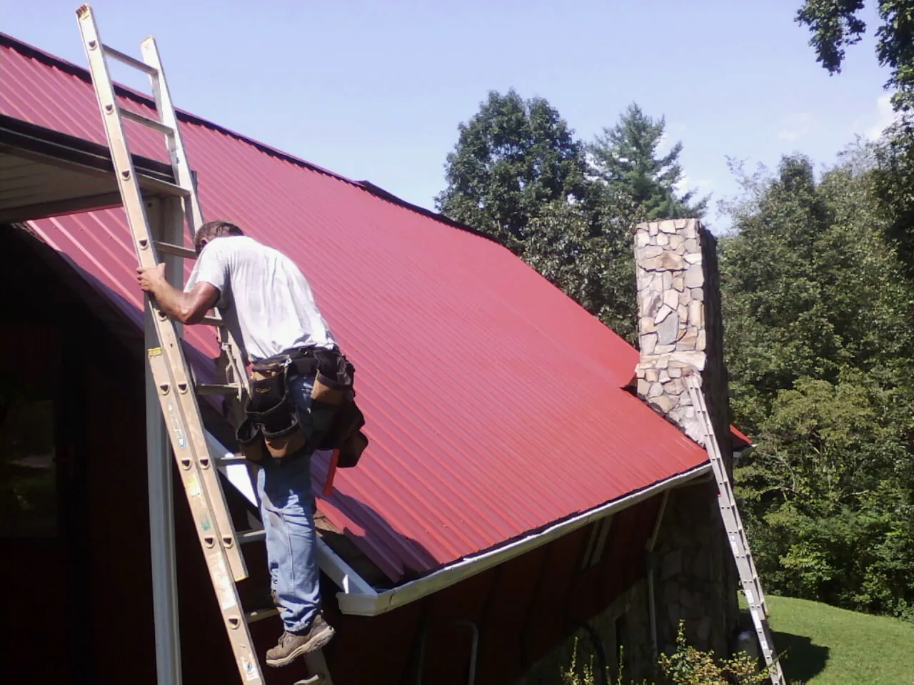 Roofer on a ladder beside a red metal roof and stone chimney — Soffit & Fascia Repair by Roofing Brothers in Starr